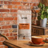 A bag of Edge Coffee Roastery El Salvador - El Muerto coffee beans stands on a wooden table in a rustic cafe setting, with a cup of latte and a potted plant in the background against a red brick wall.
