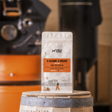 A stand-up pouch of EDGE coffee labeled 'EL SALVADOR - EL MOLINUTO' stands on top of a wooden barrel in a warm, coffee-shop-like setting with blurred coffee equipment in the background.