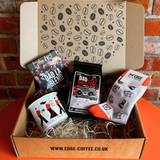 Gift box with coffee-themed items including a mug, socks, and a card, against a brick wall background.