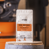 Coffee bag labeled 'EDGE' on a wooden barrel with a blurred coffee machine in the background