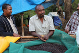 Three men sitting around a table with a green cloth covering a container of dark beans, outdoors.