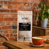 A bag of EDGE Reserve Kenya - Orange Co-Ferment coffee stands on a wooden table in a rustic cafe setting, with a brick wall background, a cup of latte, and a potted plant.