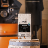 A bag of Edge Reserve Kenya - Orange CO Ferment coffee stands on a wooden barrel in a warm, rustic setting with a coffee roaster blurred in the background.