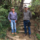 Two men standing on a dirt path with stone steps in a lush green outdoor setting. The man on the left wears a blue and white striped button-up shirt, light blue jeans, and a baseball cap.